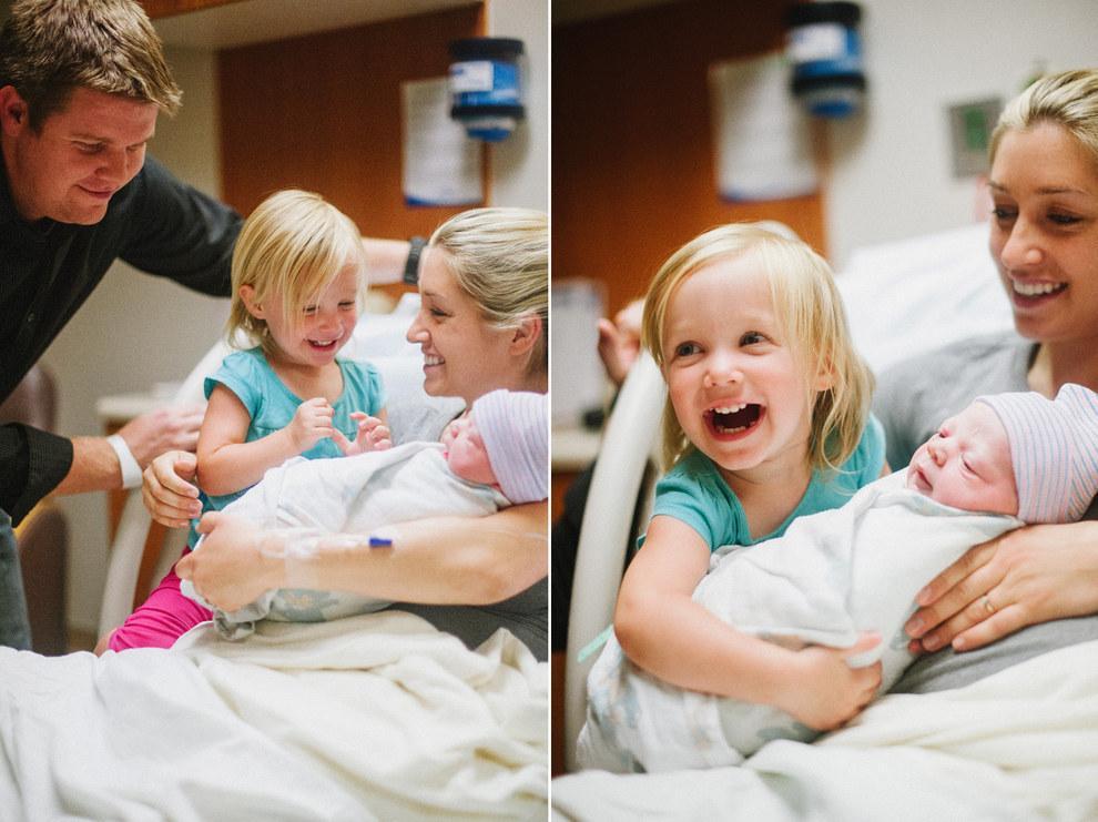 A mother holds a newborn baby in a hospital bed while a smiling young girl and a man stand nearby. The young girl looks happy and excited to meet the baby.
