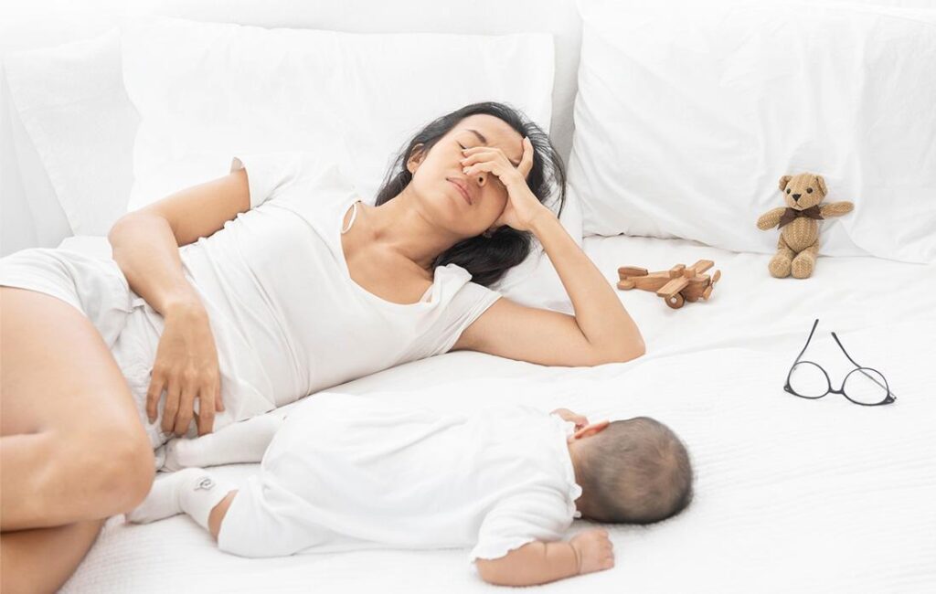 A woman lies on a bed with her eyes closed, appearing tired, while a baby lies beside her. A teddy bear, wooden toy plane, and eyeglasses are also on the bed.